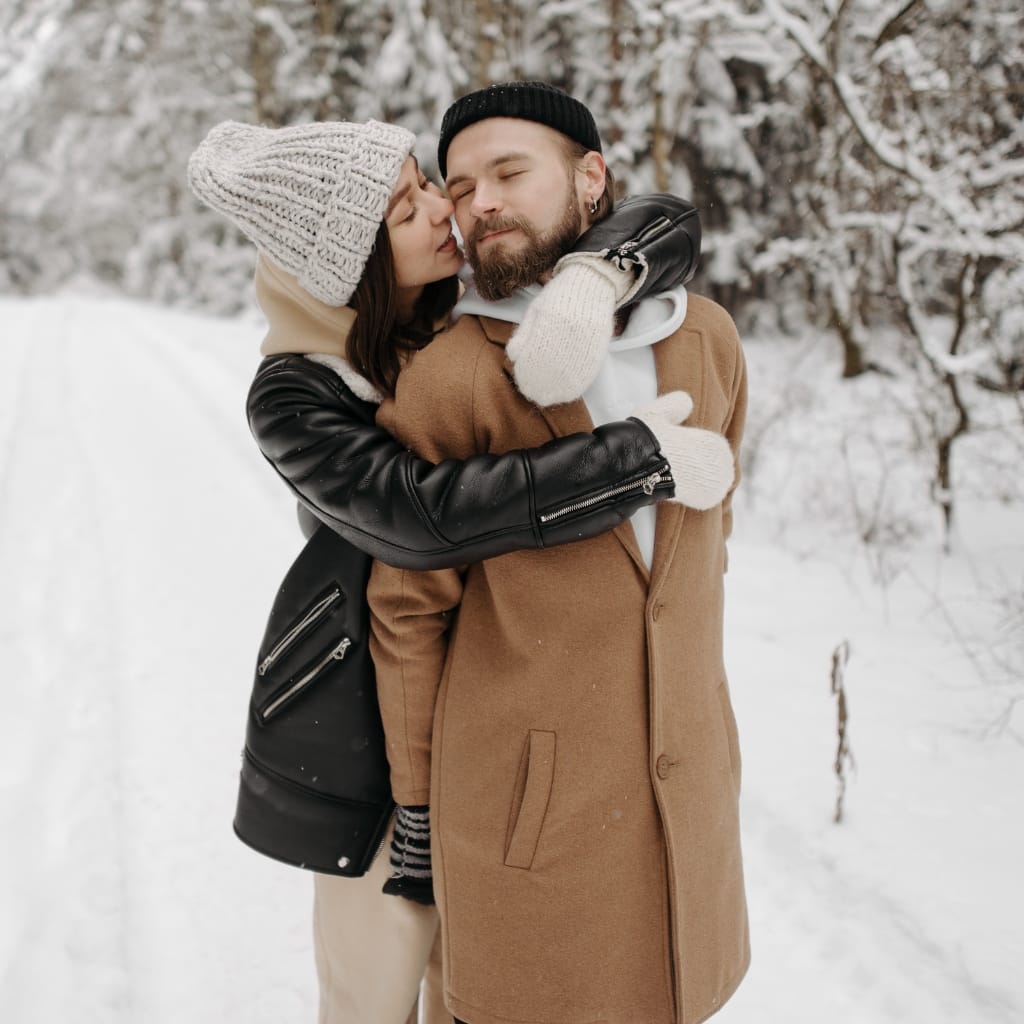 A woman and a man embrace warmly in a snowy outdoor setting. The woman is wearing a black leather jacket with a black beanie and beige gloves, while the man is dressed in a tan coat with a white hoodie, a black knit cap, and gloves. Snow-covered trees are in the background.